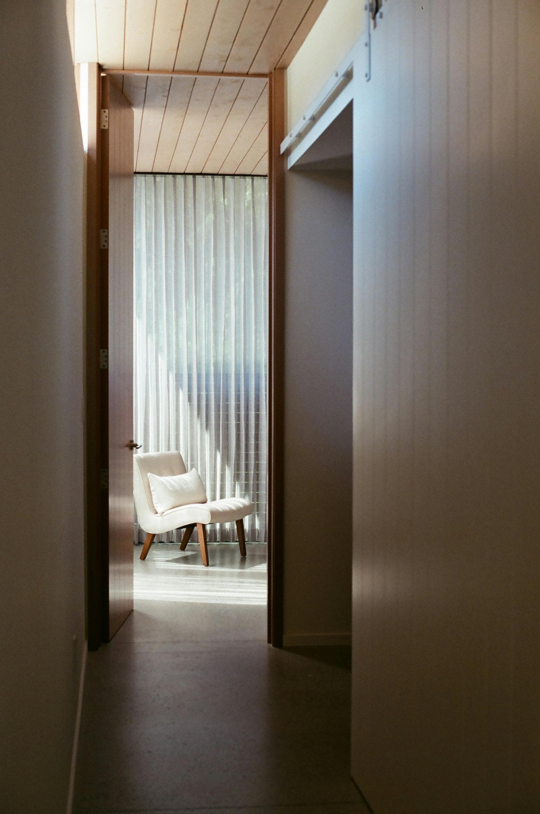 Au bout d’un couloir, une porte ouverte sur une pièce calme, un fauteuil clair devant un voilage